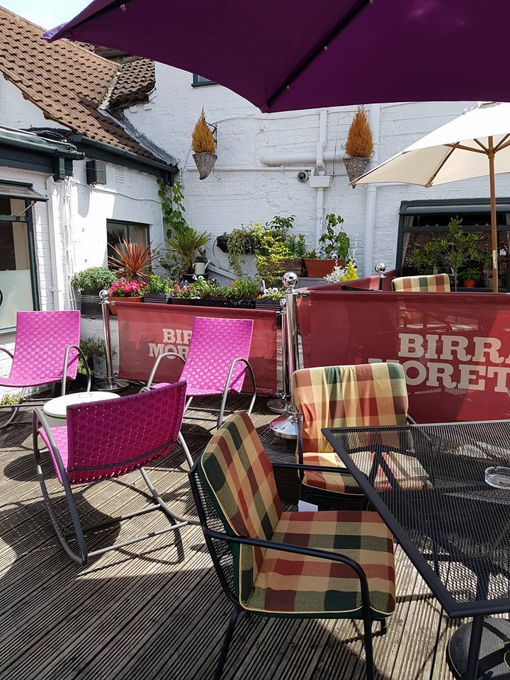 A sunny outdoor beer garden at The White Bear Pub & Restaurant in Epworth, Doncaster, featuring a mix of pink wicker and plaid cushioned chairs, dark metal tables, shade umbrellas, and decorative plants against a white building, perfect for steakhouse dining.